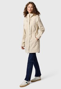 Frau mit lockigem Haar, die eine beige Kapuzenparka, dunkelblaue Jeans mit Knöchelschlitzen und weiße Sneaker mit grünen Akzenten trägt und seitlich steht.