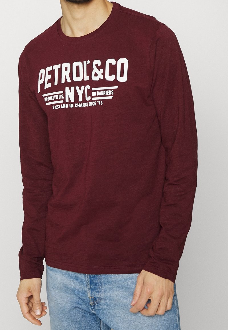 Mies, der ein kastanienfarbenes Langarmshirt mit dem weißen Aufdruck "PETROL & CO NYC" und eine blaue Jeans trägt, steht vor einem einfarbigen Hintergrund.