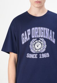 Egy sötétkék pólót viselő személy, amelyen a "GAP ORIGINAL" és a "SINCE 1969" felirat, valamint egy világos lila laurel koszorú embléma található az elején.