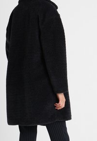 Abrigo negro oversize con un tejido texturizado y esponjoso. Cuenta con cuello y mangas largas. El diseño tiene una silueta recta.
