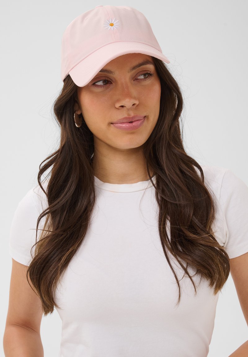 Jeune femme aux longs cheveux foncés portant une casquette rose clair avec une marguerite blanche et un t-shirt blanc, regardant légèrement sur le côté.
