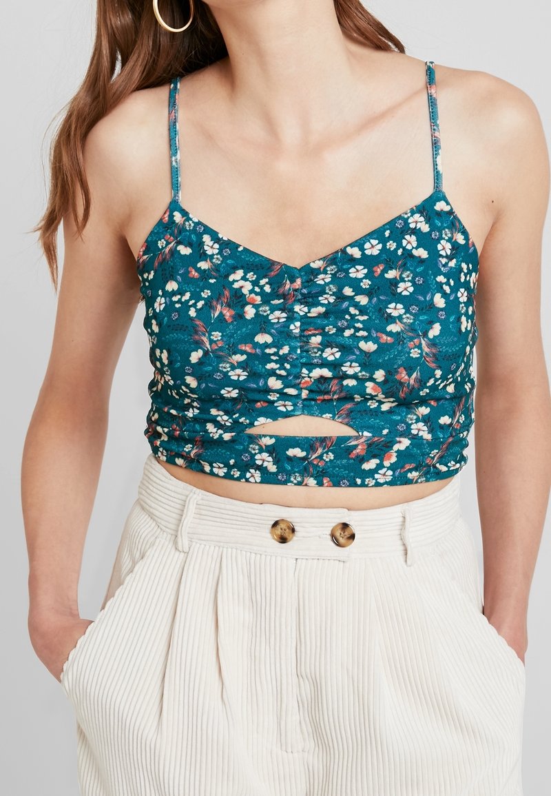Femme portant un crop top floral bleu sarcelle à fines bretelles et un pantalon beige taille haute avec des boutons, mains dans les poches, sur fond uni.