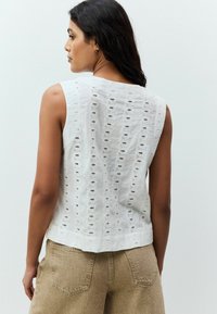 Un haut sans manches blanc présentant un motif à œillets avec des découpes. Le tissu a une finition texturée, associé à un short beige visible à la taille.