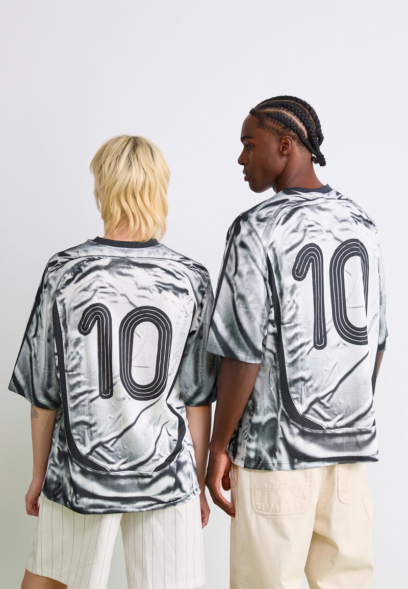 Dos personas con camisetas de tie-dye en negro y blanco a juego, con el número 10 en la parte de atrás, de espaldas frente a un fondo sencillo.