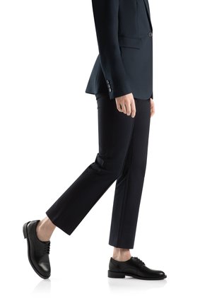 Scarpe da uomo in pelle nera con punta tonda, dettagli con lacci e una texture liscia, abbinate a pantaloni scuri su misura e a un blazer.