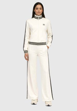 Femme debout, portant un survêtement blanc avec des bandes noires sur les manches, le pantalon et le col, assorti de baskets blanches.