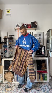 Sweater riscado com padrões castanhos, amarelos e laranjas; mangas longas e punhos canelados; formato retangular; segurado por uma pessoa com um jersey azul.