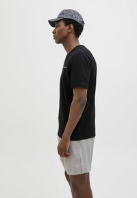 Black cotton short-sleeve shirt with a crew neck, paired with light gray shorts and a patterned dark cap, viewed from the side.