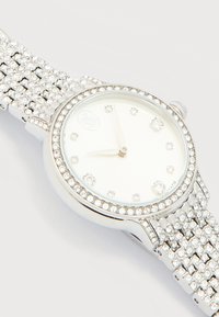 Ασημένιο ρολόι χειρός με λευκό καντράν, δείκτες ώρας από κρύσταλλο και μεταλλικό μπρασελέ και στεφάνη διακοσμημένα με κρύσταλλα, δείχνοντας ώρα 9:05.