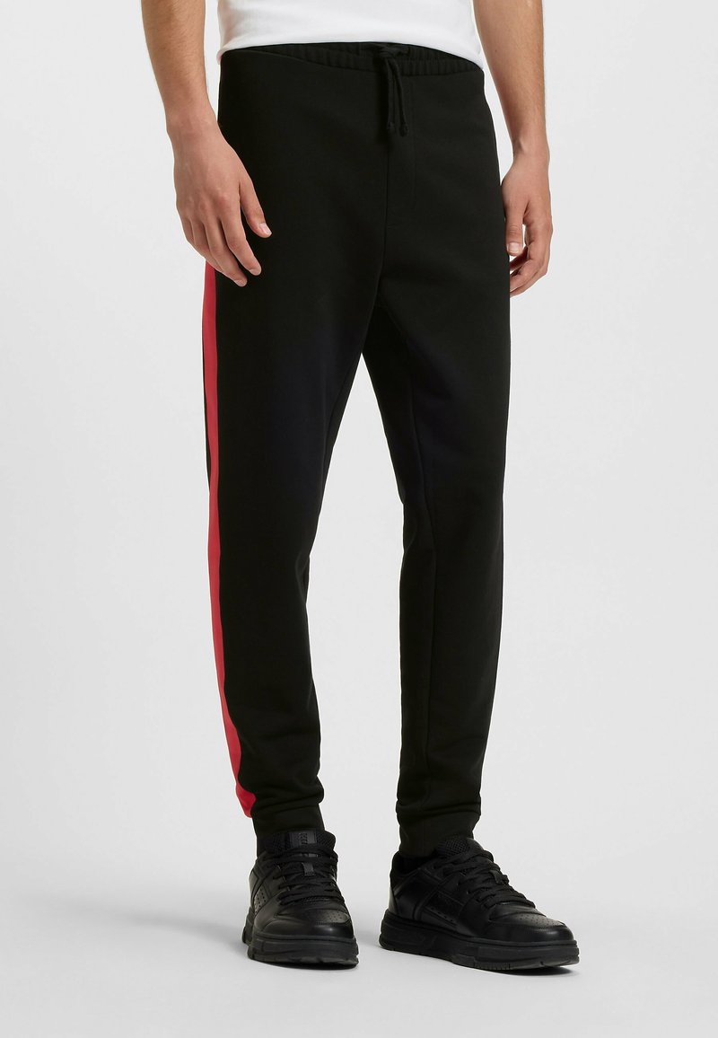 Sorte joggers med en rød side-stribe, snøre i taljen og ribbede manchetter, fremstillet af et glat, fleksibelt materiale. Velegnet til afslappet brug.