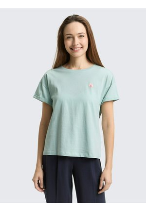 Uśmiechnięta kobieta o długich brązowych włosach, ubrana w jasnzielony krótki rękaw z małym różowym kwiatowym emblematem oraz ciemne spodnie na jasnym tle.