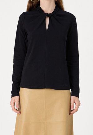 Femme portant un haut noir à manches longues avec un col noué torsadé et une jupe en cuir beige, les mains détendues le long du corps.