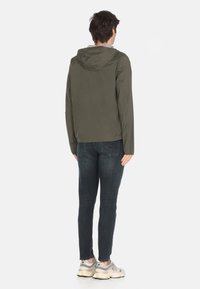 Giacca verde oliva con cappuccio, tessuto leggero, vestibilità strutturata e maniche lunghe. Abbinata a jeans scuri e sneakers.