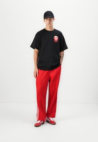 Sort t-shirt med rød grafik, røde brede bukser med hvide striber, røde sneakers med hvide detaljer og en sort cap.