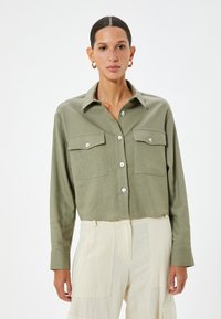 Camicia nelle tonalità di verde oliva, tagliata e con bottoni, dotata di taschini sul petto, realizzata in un tessuto leggero e con bottoni argentati. Maniche lunghe con dettagli ai polsini.