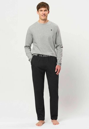 Jeune homme debout pieds nus portant un t-shirt gris à manches longues et un pantalon noir avec ceinture logo U.S. Polo sur un fond clair uni.
