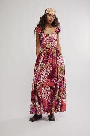 Frau mit langen lockigen Haaren, die ein maxikleid aus blumigem Patchwork, ein beiges Blumenkopftuch und dunkle Knöchelstiefel trägt, steht mit den Händen in den Taschen.