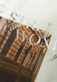 Grafický potisk s nápisem "ULICE ZÁZRAKŮ" ve stříbrném fólii na texturované látce, s ilustrací architektonické scény v teplých tónech.