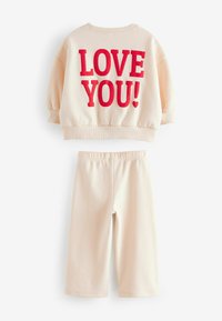 Conjunto de sudadera y pantalones en suave tela beige. La sudadera presenta un destacado texto rojo "¡TE QUIERO!". Los pantalones tienen una textura acanalada y un corte relajado.