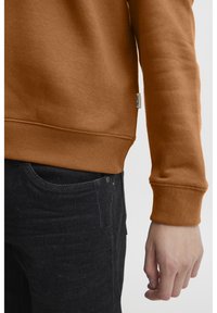 Sudadera marrón con puños y dobladillo acanalados, que presenta una pequeña etiqueta en el costado. Combinada con jeans de mezclilla oscuros. Tejido liso y texturizado.