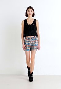 Melna adīta croptop ar saišu detaļām un daudzkrāsainiem rakstainiem šortiem; modele valkā melnus potītes zābakus uz balta fona.