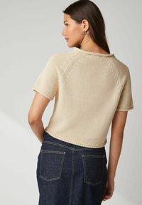 Kortärmad, krämfärgad stickad topp med rund halsringning, texturerad yta och dekorativ söm längs nederkanten, kombinerad med mörk denimkjol.