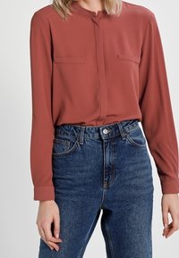 Blouse à manches longues couleur rouille avec une patte de boutonnage et deux poches poitrine, associée à un jean en denim bleu taille haute.
