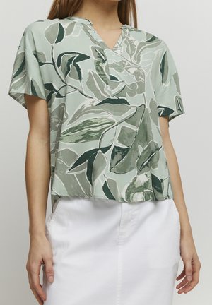 Mujer con camisa holgada de manga corta con estampado de hojas verdes y falda blanca, de pie frente a un fondo sencillo.
