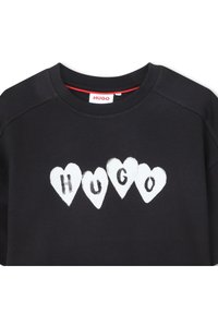 Μαύρο φούτερ με τη λέξη «HUGO» γραμμένη μέσα σε τέσσερις λευκές καρδιές στο στήθος, με κόκκινη λεπτομέρεια στο εσωτερικό του γιακά ορατή.