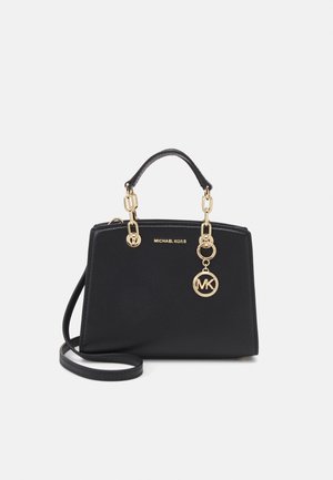 Czarna torebka Michael Kors z złotymi uchwytami na łańcuszku, zawieszką z logo MK i odpinanym paskiem na ramię na jednolitym jasnym tle.