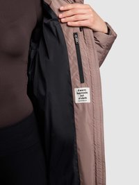 Lichtbruine pufferjas met een zwarte voering, voorzien van een zijritszak en een label met de tekst "Een warme omhelzing gemaakt van stof."