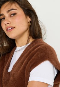 Gilet en fausse fourrure marron avec un décolleté en V, porté sur un t-shirt blanc. La texture est douce, avec des manches larges encadrant l'encolure.