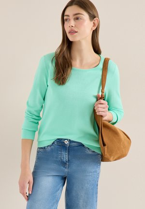 Donna che indossa una maglia a maniche lunghe verde menta e jeans blu, con una borsa a tracolla color cuoio, guarda di lato su uno sfondo neutro.