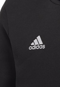 Tissu noir avec le logo Adidas blanc brodé sur la poitrine, featuring trois rayures diagonales et le nom de la marque en dessous.