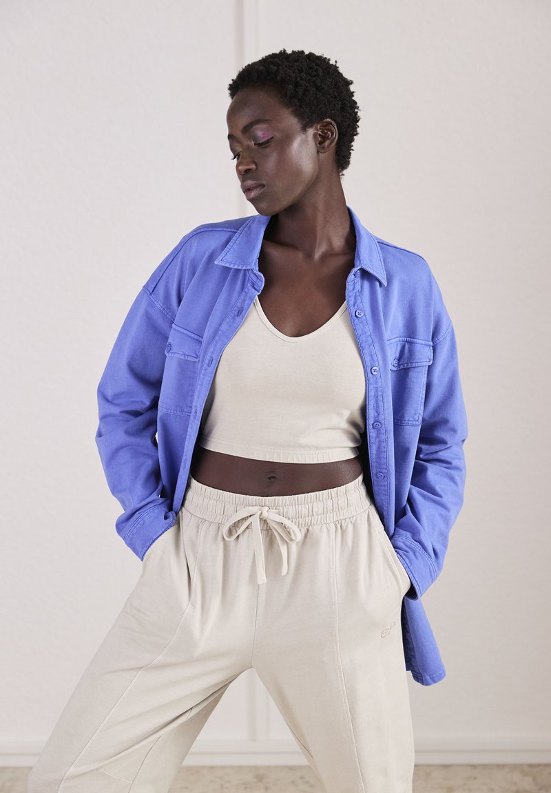 Camisa azul claro de botones, top beige sin mangas y pantalones de chándal beige con cinturilla elástica. Texturas suaves, estilo casual, corte relajado.