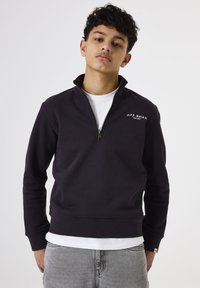 Joven con cabello rizado y oscuro que lleva una sudadera de cremallera negra con el texto "OFF ROAD", una camiseta blanca y jeans gris claro.