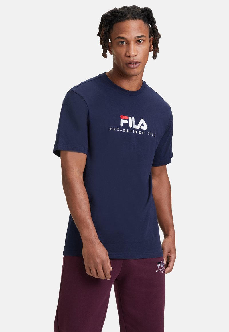 Camiseta de algodón de color azul marino con mangas cortas, que presenta un logo de Fila bordado en rojo y blanco y el texto "ESTABLECIDO 1911" en el frente.
