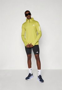 Green long-sleeve sports top with half-zip and white Adidas stripes, paired with black shorts. Model wears sunglasses and athletic shoes.