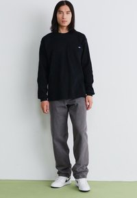 Μαύρο μπλουζάκι με μακριά μανίκια και τσέπη, συνδυασμένο με γκρι παντελόνι και λευκά αθλητικά παπούτσια. Απλός σχεδιασμός, λεία υφή, ελάχιστη σήμανση.