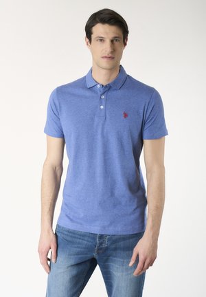 Giovane uomo che indossa una polo blu a maniche corte con logo rosso e jeans blu sbiaditi, in piedi contro uno sfondo chiaro e semplice.