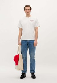 T-shirt en coton blanc avec logo rouge, jeans en denim bleu, chaussures noires et un sac fourre-tout en toile naturelle avec une casquette rouge glissée à l'intérieur. Design simple.
