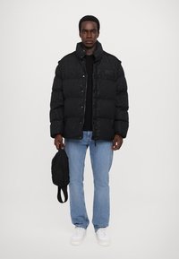 Black puffer jacket with a shiny finish, zip and snap closure, side pockets, worn over a black shirt, paired with light blue jeans and white sneakers.