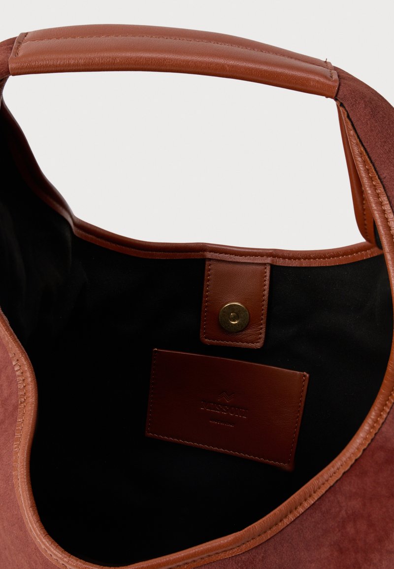 Sac fourre-tout en daim marron avec une poigne en cuir, fermeture à pression dorée et doublure intérieure noire. Présente une étiquette de marque en relief.