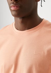 Camiseta de algodón color durazno con cuello redondo y logo de Nike bordado en el pecho, que presenta una textura suave y lisa.