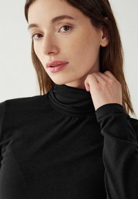 Melnas turtlenecks, kas izgatavots no mīkstas, elastīgas auduma, ar garām piedurknēm un pieguļošiem aproču galiem, kā arī sakļautu dekoltē. Vienkārša un gluda tekstūra.