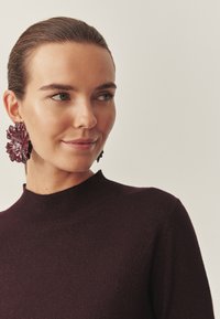 Pull en maille bordeaux à col montant associé à de grandes boucles d'oreilles florales bordeaux, surface texturée, et vue rapprochée du visage.