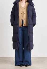 Granatowa pikowana kurtka puchowa z kapturem, długiego kroju. Zestawiona beżowym dzianinowym kardiganem i szerokimi niebieskimi dżinsami, widoczne czarne buty.