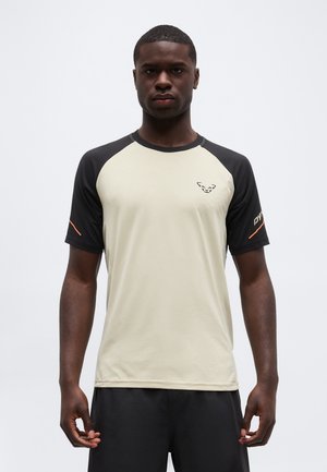 Noor mees, kes kannab beeži ja musta lühikeste varrukatega spordisärki rinnal logoga ja musti lühikesi pükse, seisab ühevärvilise tausta ees.