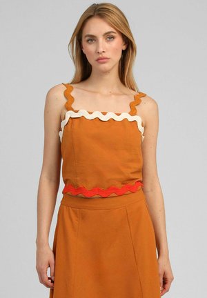 Femme portant un crop top orange sans manches avec un bord ondulé blanc et rouge, associé à une jupe taille haute orange sur fond gris uni.
