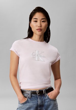 Mujer con cabello oscuro que lleva una camiseta rosa claro de Calvin Klein y vaqueros azules con las manos en los bolsillos frente a un fondo gris liso.